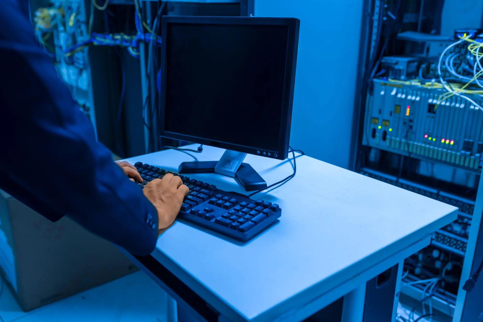 IT technician typing at a terminal beside network racks in a blue-lit server room