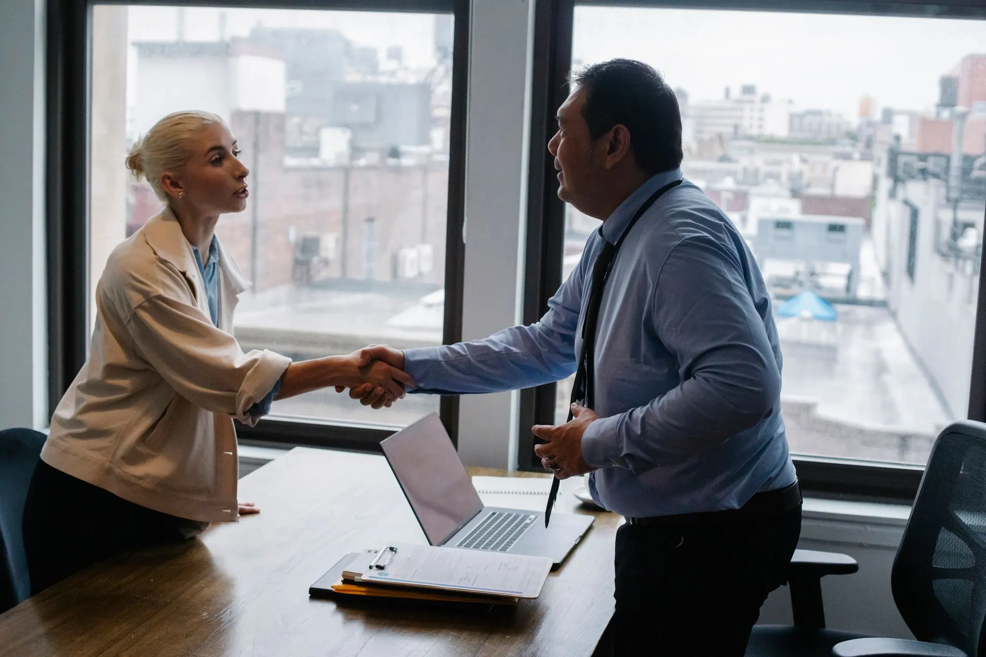 Two professionals shaking hands across a desk by a bright window