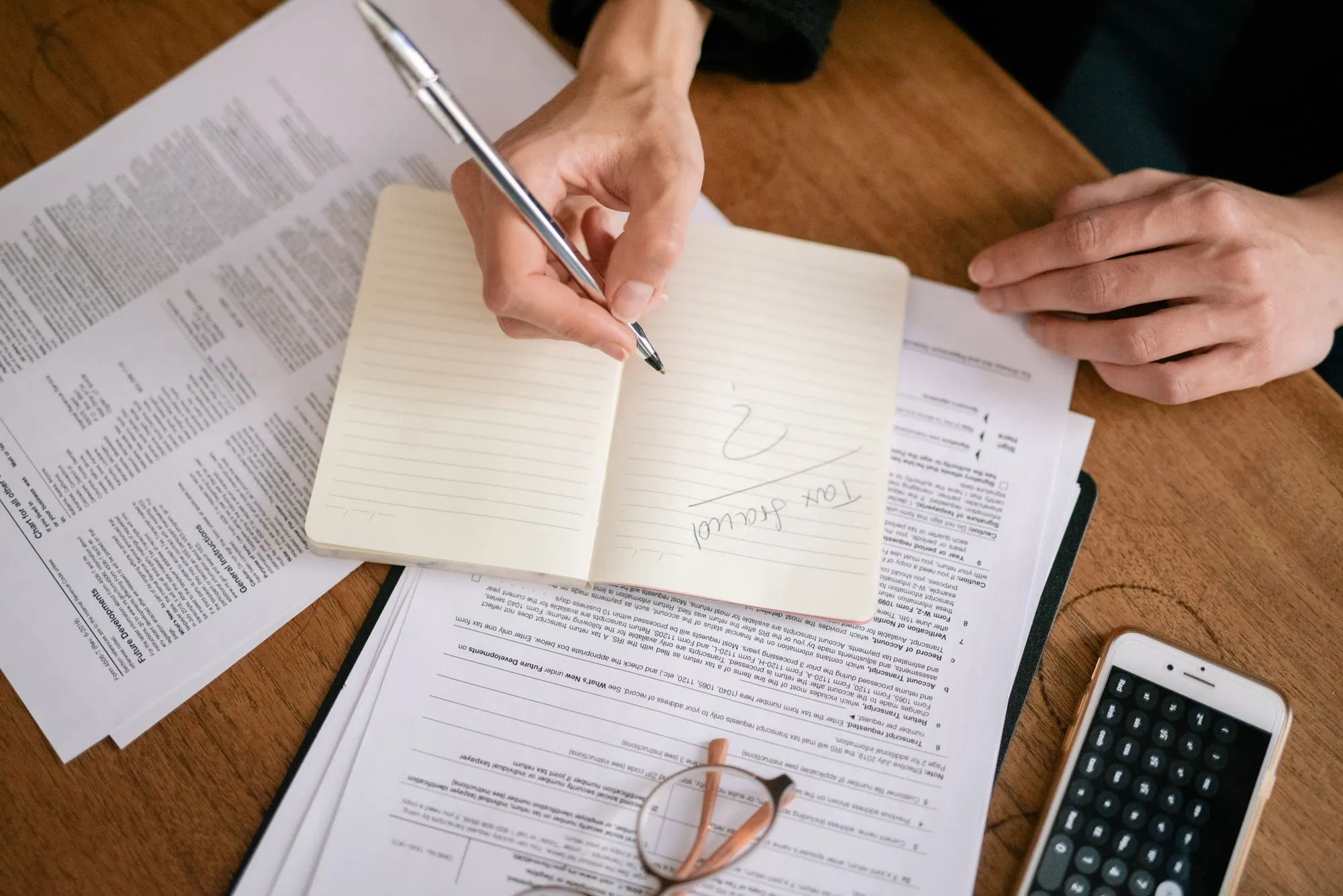 Hands writing notes in a notebook beside paperwork and a smartphone